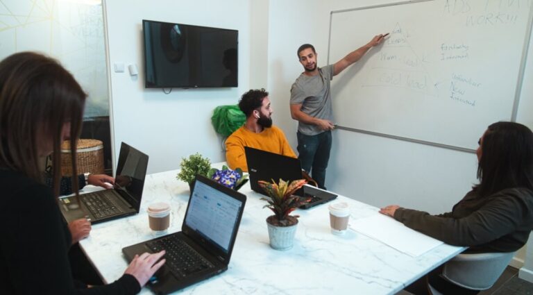 Team members sit at a desk on their laptops whilst a man presents at a whiteboard
