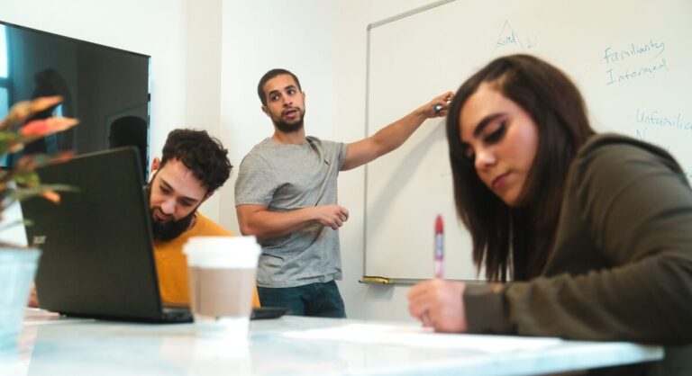 Two team members sitting at a desk writing whilst a man is standing talking at a whiteboard